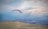 Paragliders soar over a vast, open landscape with rolling hills and a dramatic sky filled with clouds and sunlight rays.