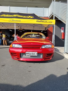 A red sports car is parked in front of a yellow canopy tent with 'Haltech' branding. Several people stand nearby, and the car is positioned on a gray asphalt surface. Metal structures and a building are visible in the background.