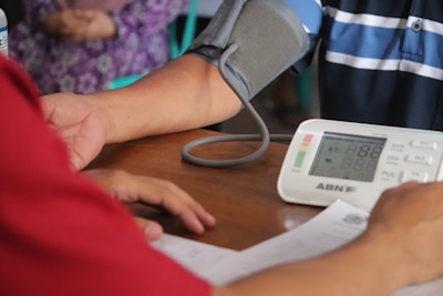 A person is getting their blood pressure checked with an electronic monitor. A cuff is wrapped around the upper arm and connected to the device on a table. Another person is seen sitting opposite, possibly aiding the process.