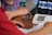 A person is getting their blood pressure checked with an electronic monitor. A cuff is wrapped around the upper arm and connected to the device on a table. Another person is seen sitting opposite, possibly aiding the process.