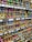 Numerous cardboard boxes neatly arranged on shelves, each containing multiple brightly colored spray paint cans, are visible. The cans are organized by color, including shades of green, pink, blue, yellow, and more. Labels with prices and item details are affixed to the shelf edges.