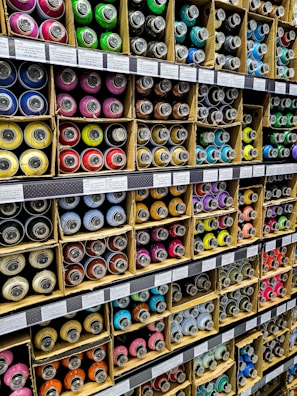 Numerous cardboard boxes neatly arranged on shelves, each containing multiple brightly colored spray paint cans, are visible. The cans are organized by color, including shades of green, pink, blue, yellow, and more. Labels with prices and item details are affixed to the shelf edges.
