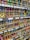 Numerous cardboard boxes neatly arranged on shelves, each containing multiple brightly colored spray paint cans, are visible. The cans are organized by color, including shades of green, pink, blue, yellow, and more. Labels with prices and item details are affixed to the shelf edges.