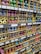 Numerous cardboard boxes neatly arranged on shelves, each containing multiple brightly colored spray paint cans, are visible. The cans are organized by color, including shades of green, pink, blue, yellow, and more. Labels with prices and item details are affixed to the shelf edges.