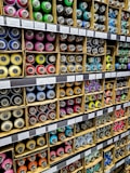 Numerous cardboard boxes neatly arranged on shelves, each containing multiple brightly colored spray paint cans, are visible. The cans are organized by color, including shades of green, pink, blue, yellow, and more. Labels with prices and item details are affixed to the shelf edges.