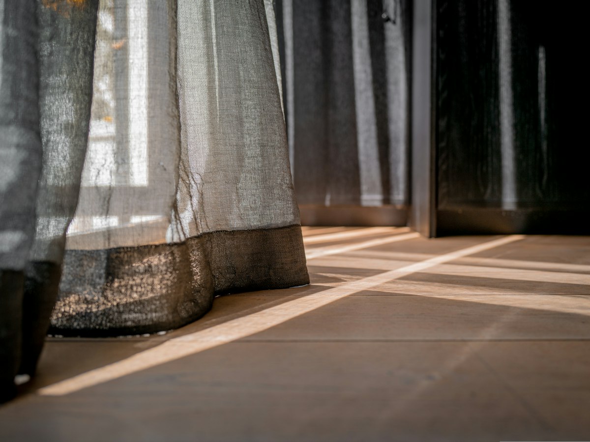 Cortinas de gasa de lino gris que llegan al piso de madera, con rayos de sol que entran por la ventana y se reflejan en el suelo