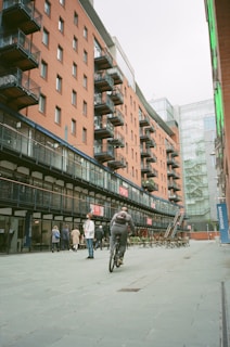 A cyclist rides along a wide pedestrian path adjacent to a series of modern brick apartment buildings with numerous balconies. Several people are walking on the path, and there is outdoor seating arranged nearby. The atmosphere is urban and lively, with modern architecture featuring prominently.