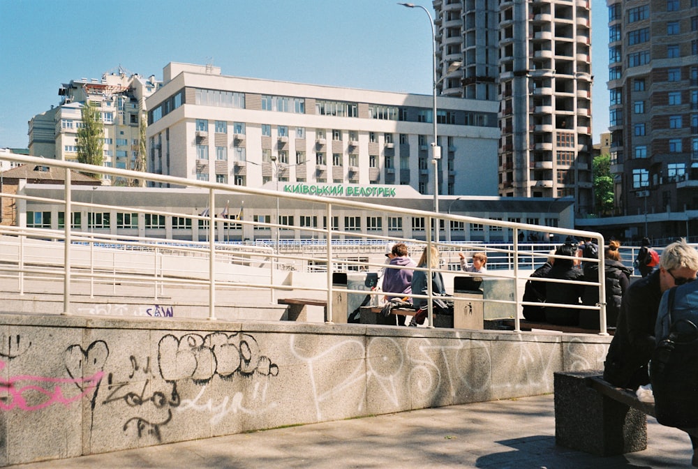 昼間、白いコンクリートの建物の近くのベンチに座っている2人の男性