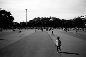 A wide, open public square filled with numerous people engaged in various activities. Some individuals are walking, while others are playing or relaxing. Children can be seen playing with a ball. Tall trees and a streetlight are visible in the background under a clear sky.