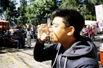 A young person wearing a hoodie drinks from a clear glass. The scene takes place outdoors where there are people in the background, some wearing jackets, and the presence of motorcycles indicates an urban setting. Sunlight creates shadows, adding contrast to the image.
