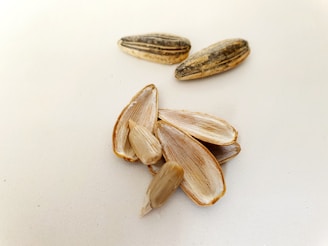 Several sunflower seeds are scattered on a light surface. Some of the seeds are whole, while others are shelled, revealing their inner kernels. The shells are elongated and striped with a mixture of dark and light brown colors.