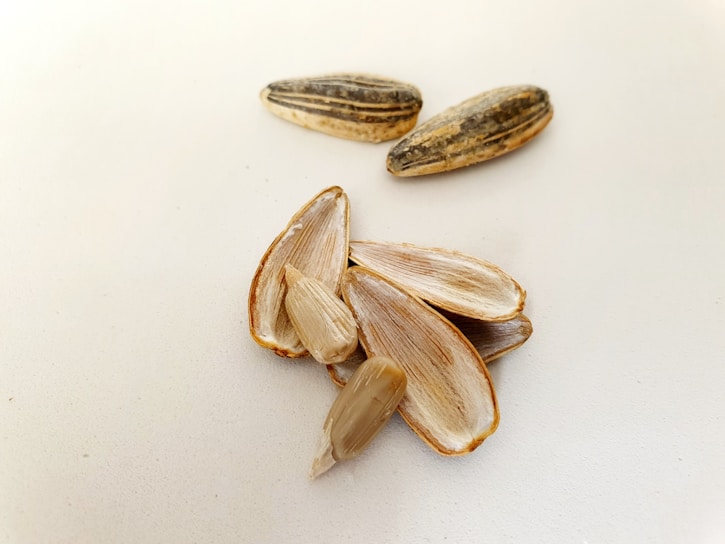 Several sunflower seeds are scattered on a light surface. Some of the seeds are whole, while others are shelled, revealing their inner kernels. The shells are elongated and striped with a mixture of dark and light brown colors.