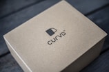 A brown cardboard box is placed on a dark wooden surface. The box features a simple design with a logo of a cup and the brand name 'CURVD' printed in black on its top surface.