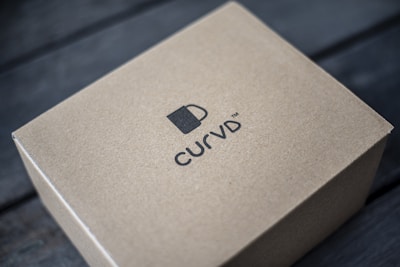A brown cardboard box is placed on a dark wooden surface. The box features a simple design with a logo of a cup and the brand name 'CURVD' printed in black on its top surface.