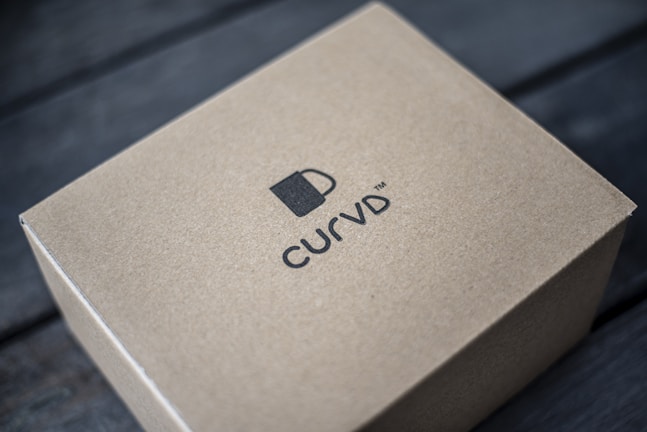 A brown cardboard box is placed on a dark wooden surface. The box features a simple design with a logo of a cup and the brand name 'CURVD' printed in black on its top surface.