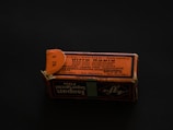 A rectangular cardboard box with a worn, orange-and-black printed label displaying text in multiple languages. The label includes technical specifications, possibly related to a photographic product or film. The box is set on a dark background, creating a contrast that highlights its aged appearance.