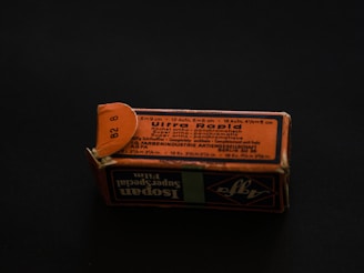 A rectangular cardboard box with a worn, orange-and-black printed label displaying text in multiple languages. The label includes technical specifications, possibly related to a photographic product or film. The box is set on a dark background, creating a contrast that highlights its aged appearance.