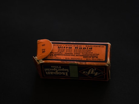 A rectangular cardboard box with a worn, orange-and-black printed label displaying text in multiple languages. The label includes technical specifications, possibly related to a photographic product or film. The box is set on a dark background, creating a contrast that highlights its aged appearance.