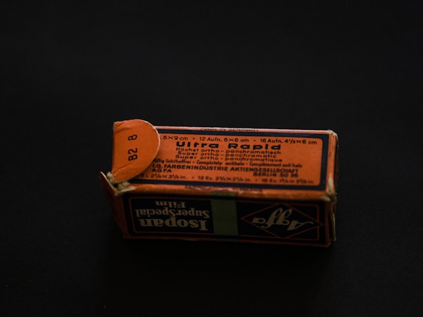 A rectangular cardboard box with a worn, orange-and-black printed label displaying text in multiple languages. The label includes technical specifications, possibly related to a photographic product or film. The box is set on a dark background, creating a contrast that highlights its aged appearance.