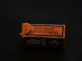 A rectangular cardboard box with a worn, orange-and-black printed label displaying text in multiple languages. The label includes technical specifications, possibly related to a photographic product or film. The box is set on a dark background, creating a contrast that highlights its aged appearance.