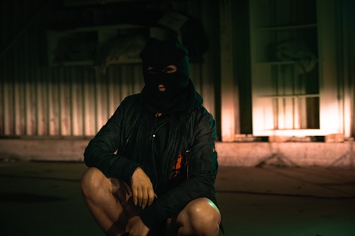 A person wearing a black ski mask and dark clothing crouches indoors, with a dimly lit and industrial-style background featuring a corrugated metal wall.