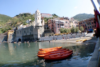 A scenic coastal town features charming buildings of varying pastel colors situated on a hillside. In the foreground, bright orange kayaks float on the calm sea along a sandy beach where people are swimming and sunbathing. A historic stone clock tower adds a picturesque element to the scene.