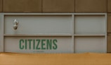 A security camera is mounted on a building wall above a panel with the text 'CITIZENS OF CHELSEA' printed in green letters. The structure has a beige exterior and a yellowish tarp or awning partially covers the lower portion.