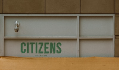 A security camera is mounted on a building wall above a panel with the text 'CITIZENS OF CHELSEA' printed in green letters. The structure has a beige exterior and a yellowish tarp or awning partially covers the lower portion.