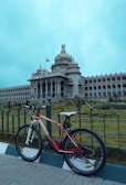 A red bicycle is parked in front of an ornate and expansive government building with a large dome and classical columns. The building features intricate architectural details and an Indian flag flying high. The sky is overcast, lending a moody atmosphere to the scene.