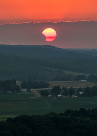 A partially eclipsed sun hovers over a vast landscape with rolling hills, farmlands, and scattered trees. The sky is painted in vibrant shades of orange and red as the sun approaches the horizon. Below, rural fields and clusters of farm buildings are visible in diminishing light, adding a serene touch to the scene.
