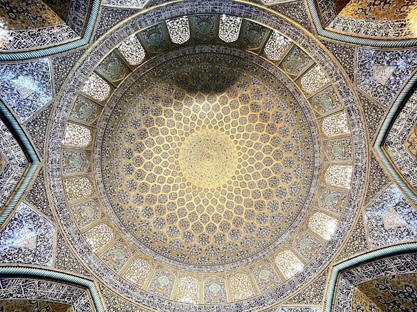 A beautifully intricate dome interior featuring detailed geometric patterns and calligraphy. The design employs a harmonious blend of blue, gold, and white colors, creating a mesmerizing visual effect. The symmetry and craftsmanship reflect traditional Islamic architecture.