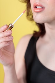 A close-up view of a person applying peach-colored lip gloss with a wand applicator. The person is wearing a black top, and the background is bright yellow, highlighting the warm tones of the lips and nail polish.