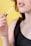 A close-up view of a person applying peach-colored lip gloss with a wand applicator. The person is wearing a black top, and the background is bright yellow, highlighting the warm tones of the lips and nail polish.