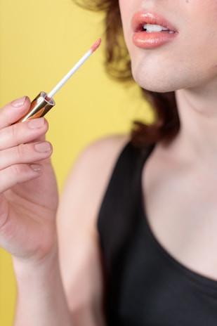 A close-up view of a person applying peach-colored lip gloss with a wand applicator. The person is wearing a black top, and the background is bright yellow, highlighting the warm tones of the lips and nail polish.