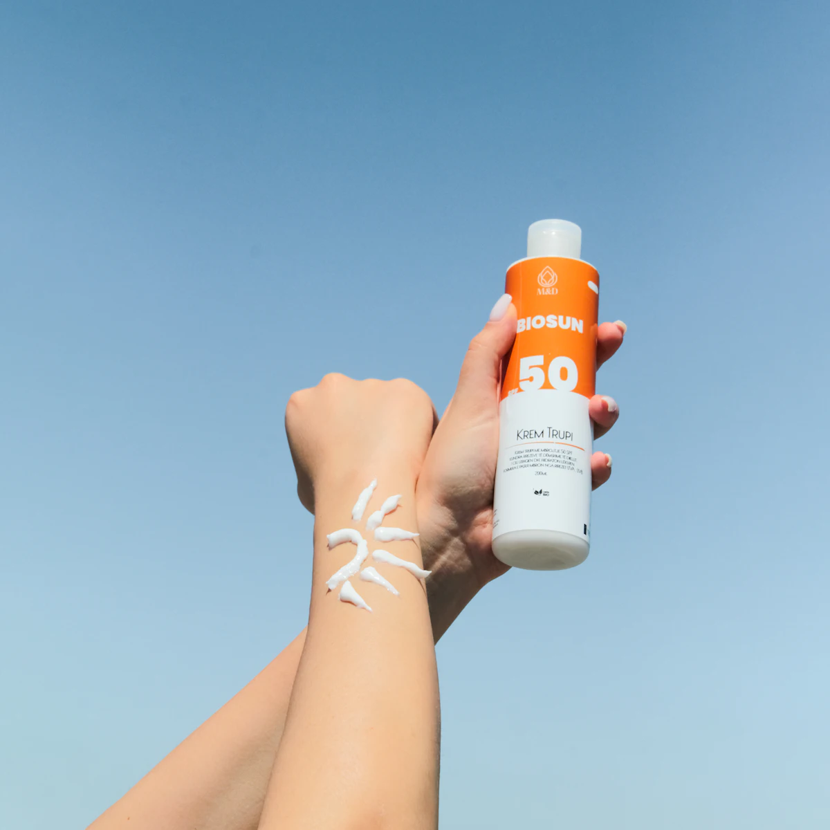 Tangan memegang botol tabir surya SPF 50 di bawah langit biru cerah sebagai perlindungan kulit dari paparan matahari