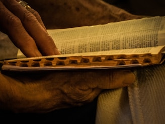 A close-up view of an open book with well-worn pages being held by a person's hands. The individual's fingers are partially visible, gently turning a page. The lighting has a warm tone, casting a soft glow on the book and hands, suggesting an intimate and serene atmosphere.