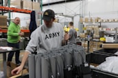 Several people are working in a warehouse setting, with shelves, boxes, and electronic components visible. One person in the foreground is wearing a sweatshirt with 'ARMY' on it, and appears to be handling cables or devices. Another person in a green hoodie stands nearby, looking on.