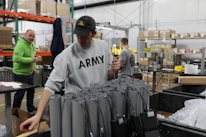 Several people are working in a warehouse setting, with shelves, boxes, and electronic components visible. One person in the foreground is wearing a sweatshirt with 'ARMY' on it, and appears to be handling cables or devices. Another person in a green hoodie stands nearby, looking on.