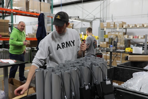 Several people are working in a warehouse setting, with shelves, boxes, and electronic components visible. One person in the foreground is wearing a sweatshirt with 'ARMY' on it, and appears to be handling cables or devices. Another person in a green hoodie stands nearby, looking on.