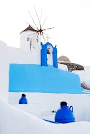 A traditional windmill with a thatched roof stands prominently against a clear sky, surrounded by iconic white and blue architecture typical of the Mediterranean. Bright blue domes and structures contrast with the stark white walls, capturing the aesthetic of a coastal town. A bell hangs beneath a blue archway, symbolizing local culture.