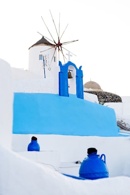 A traditional windmill with a thatched roof stands prominently against a clear sky, surrounded by iconic white and blue architecture typical of the Mediterranean. Bright blue domes and structures contrast with the stark white walls, capturing the aesthetic of a coastal town. A bell hangs beneath a blue archway, symbolizing local culture.