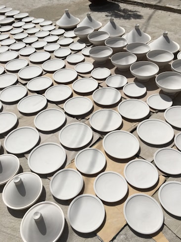 Several unglazed clay tajine pots are arranged neatly on a cardboard surface outdoors. The pots, consisting of round bases and conical lids, are organized in rows, casting subtle shadows on the ground. The sunlight creates a soft interplay of light and shadow across the surface of the pots.