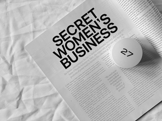 A monochromatic photograph features an open magazine or booklet with the headline 'SECRET WOMEN'S BUSINESS' in bold black letters. The text below the headline discusses female entrepreneurs and the economic recovery post-COVID-19. A round white object with a black symbol partially obscures part of the text. The background appears to be a crumpled white fabric.