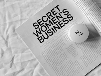 A monochromatic photograph features an open magazine or booklet with the headline 'SECRET WOMEN'S BUSINESS' in bold black letters. The text below the headline discusses female entrepreneurs and the economic recovery post-COVID-19. A round white object with a black symbol partially obscures part of the text. The background appears to be a crumpled white fabric.