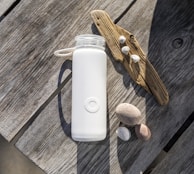 A white water bottle with a handle is placed on a weathered wooden table. Nearby, a piece of driftwood is adorned with small seashells. Several smooth stones are scattered at the side, creating a natural beach-like arrangement.