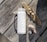 A white water bottle with a handle is placed on a weathered wooden table. Nearby, a piece of driftwood is adorned with small seashells. Several smooth stones are scattered at the side, creating a natural beach-like arrangement.