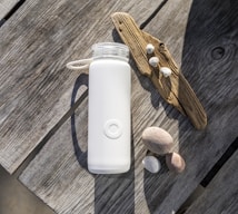 A white water bottle with a handle is placed on a weathered wooden table. Nearby, a piece of driftwood is adorned with small seashells. Several smooth stones are scattered at the side, creating a natural beach-like arrangement.