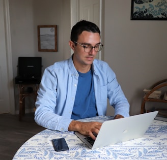 A person wearing glasses is working on a laptop at a round table covered with a blue and white patterned tablecloth. There is a smartphone placed on the table. The room has plain walls with a framed picture and a piece of art hanging. A piece of furniture with shelves is visible in the background.