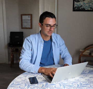A person wearing glasses is working on a laptop at a round table covered with a blue and white patterned tablecloth. There is a smartphone placed on the table. The room has plain walls with a framed picture and a piece of art hanging. A piece of furniture with shelves is visible in the background.