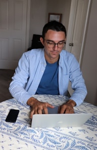 Photo of a man working on a laptop with eyeglasses beside him.